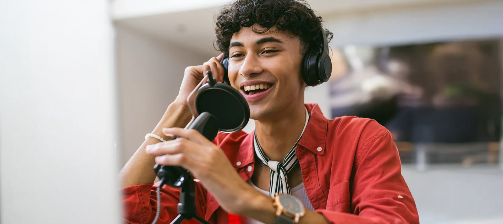A young male voice actor with curly hair wearing headphones and smiling while speaking into a studio microphone with a pop filter, representing audio content creation